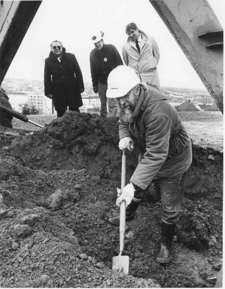 Black and white photo of Lou Collins digging with a shovel beneath an excavator, with City officials looking on.