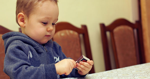 Photograph of a young boy sitting at a dining table and playing with a box of matches. He is trying to light a wooden match against the side of the box of matches.