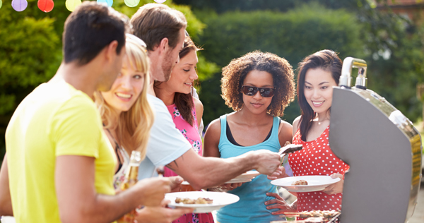 Close up photograph of several young adults wearing colorful summer clothing gathered around a gas barbecue while one man is placing grilled food on their plates
