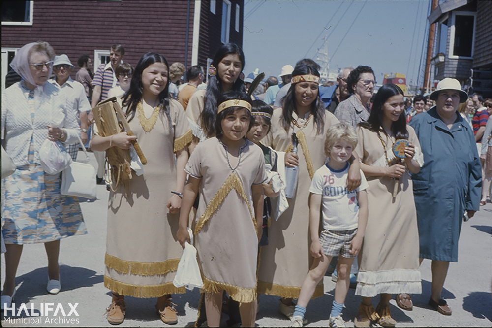 Image of a group of Mi'kmaq people in traditional dress posing with a family during Natal Day celebrations on the Halifax waterfront