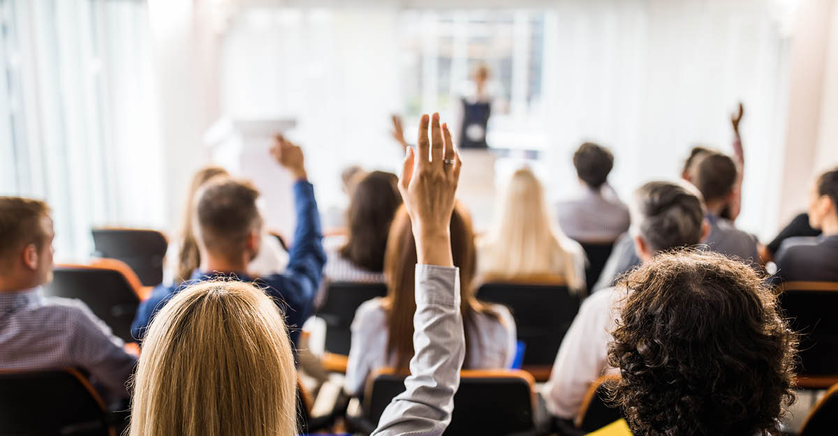 Group of people sitting, facing a white board, with their hands raised