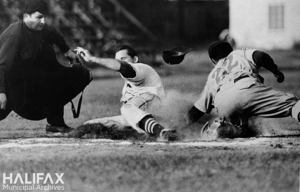 Black and white photograph of a player sliding into home plate while the catcher attempts to tag and the umpire watches to call the play