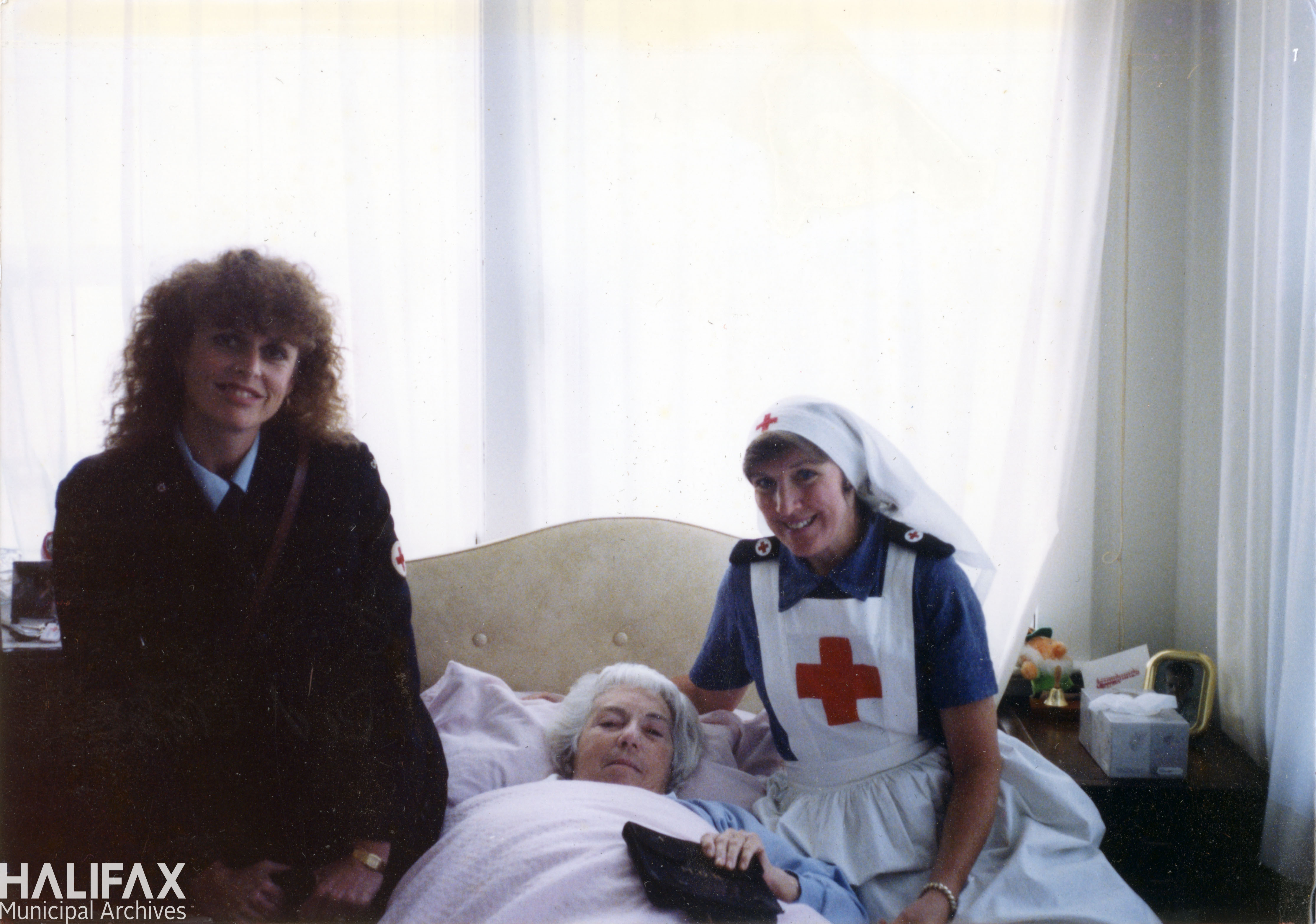 Colour image of 2 nurses on either side of elderly woman lying in a bed in front of a window.