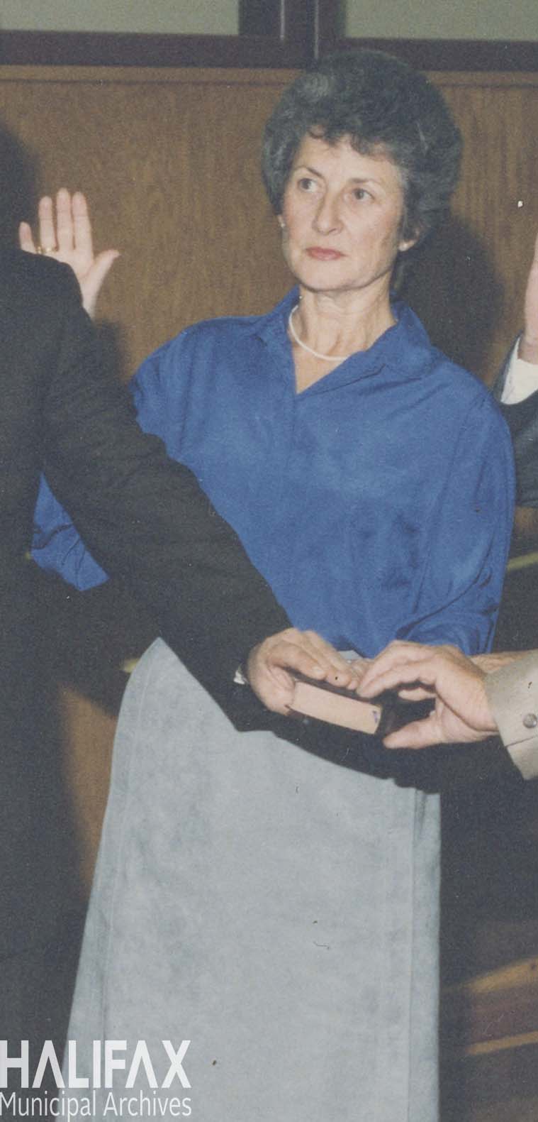 Colour photograph of a dark haired woman wearing a blue blouse and grey skirt with her right hand raised and left hand on a bible