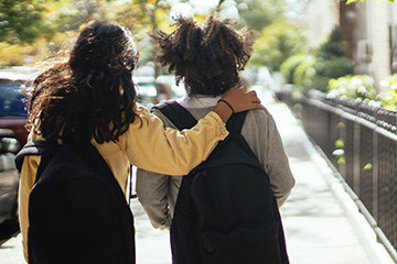 Two young female students walk along the sidewalk with black bookbags on. The girl on the right has her right arm wrapped around her friend. They are facing away from the camera.