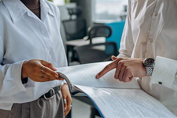 An image shows a zoomed in shot of two people, one who is holding an open document, and a second person pointing to something on a page.