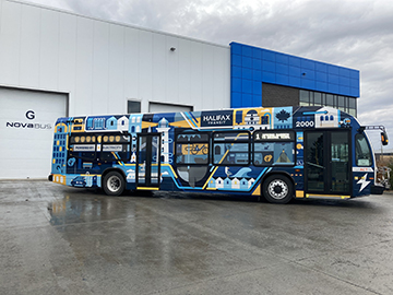 A photo of Halifax Transit’s pilot electric bus parked outside a bus garage. The bus is white on the front, like other Halifax Transit buses, but is wrapped in a special design on the back and sides. The design includes drawings of lighthouses, buses, ferries, the Citadel Hill clock tower, the Wave statue, seagulls, whales, lighting bolts, and more all connected by lines hinting at electric cables.