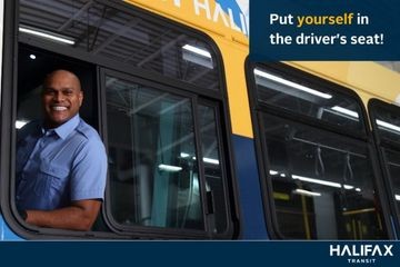 A bus driver sits in the driver seat of the bus. He is looking out the window and smiling. 